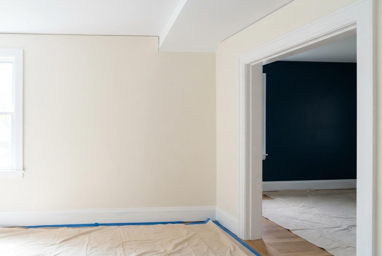 Bright cream room under renovation with blue painter’s tape, floor coverings, and a doorway to a dark blue room.