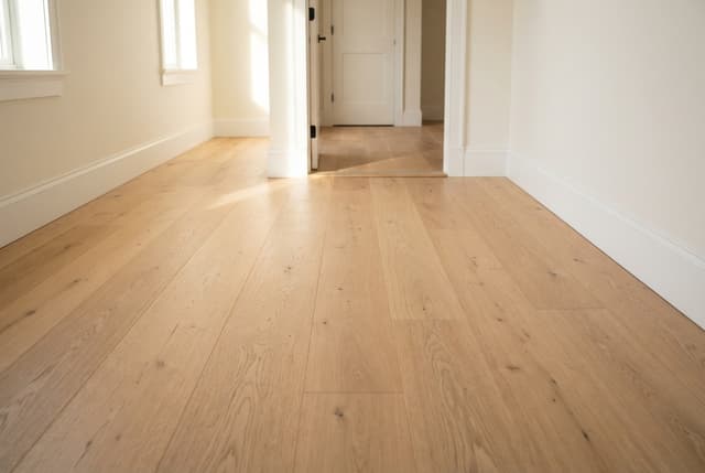 Empty sunlit room with light wood floors, white walls, windows, and open doors.