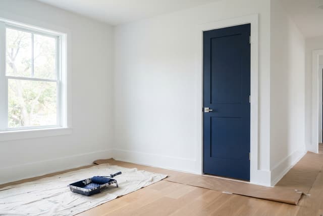 Bright white room under renovation with a navy door, window, covered wood floor, drop cloth, and paint tray.