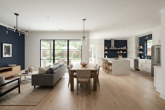 Bright open-plan living room, dining area, and navy-and-white kitchen with a backyard visible through glass doors.
